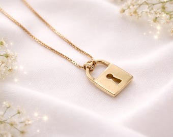 May include: A gold-colored padlock pendant necklace. The pendant is a small, square-shaped padlock with a keyhole. The necklace chain is a delicate gold color. The background is a white, silky fabric with small white flowers.