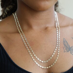 Puede incluir: Dos delicados collares dorados en capas. Cada collar presenta una serie de peque&ntilde;os eslabones ovalados alargados. Los collares se usan sobre una parte superior oscura, creando un contraste que resalta el dise&ntilde;o de la joyer&iacute;a.