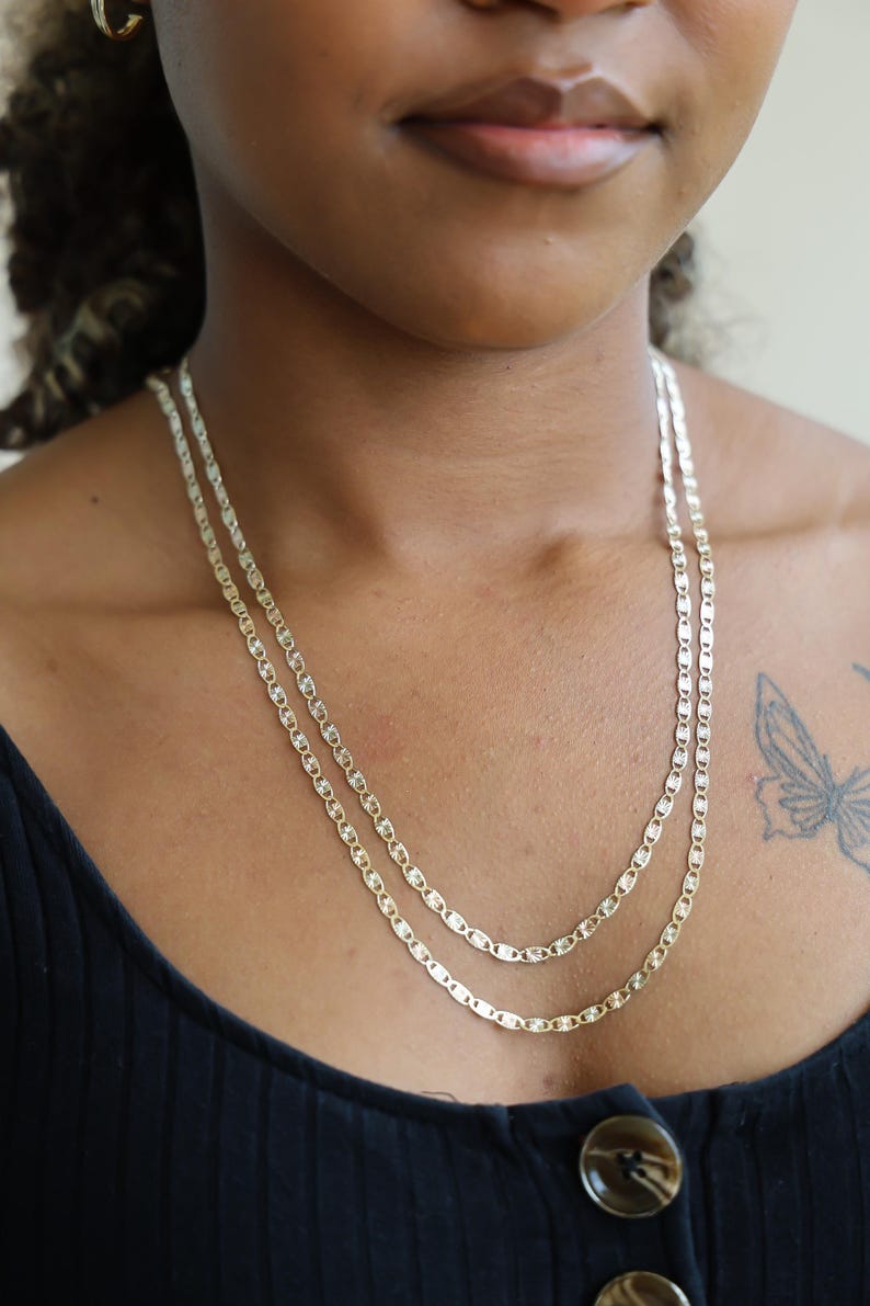 Puede incluir: Dos delicados collares dorados en capas. Cada collar presenta una serie de peque&ntilde;os eslabones ovalados alargados. Los collares se usan sobre una parte superior oscura, creando un contraste que resalta el dise&ntilde;o de la joyer&iacute;a.