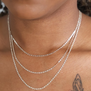 Puede incluir: Primer plano de tres delicados collares de cadena plateada de diferentes longitudes. Las cadenas est&aacute;n superpuestas, creando un look elegante. El tono de piel es medio y un tatuaje de mariposa es visible en el hombro.