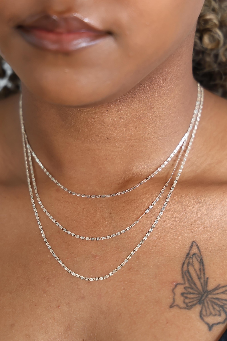 Puede incluir: Primer plano de tres delicados collares de cadena plateada de diferentes longitudes. Las cadenas est&aacute;n superpuestas, creando un look elegante. El tono de piel es medio y un tatuaje de mariposa es visible en el hombro.