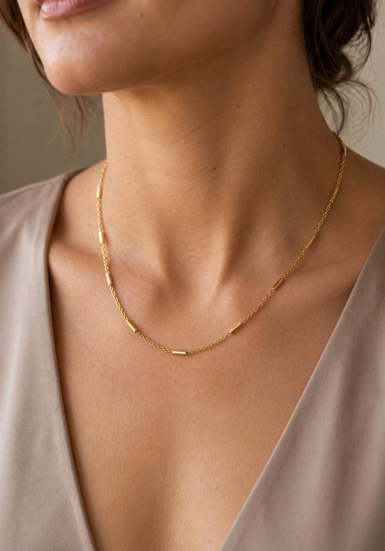 Puede incluir: Un collar de cadena dorada que presenta peque&ntilde;os acentos de barras met&aacute;licas cil&iacute;ndricas espaciadas uniformemente a lo largo de la delicada cadena de eslabones. El collar descansa sobre el cuello, mostrando un dise&ntilde;o simple y pulido.