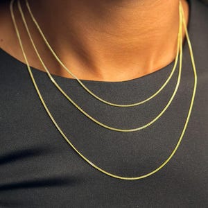 May include: Three layered gold-tone chains with a delicate curb link design. The chains are worn around the neck, featuring a consistent 1.2mm width. The metallic finish is bright and reflective, creating a classic layered look against a dark fabric.