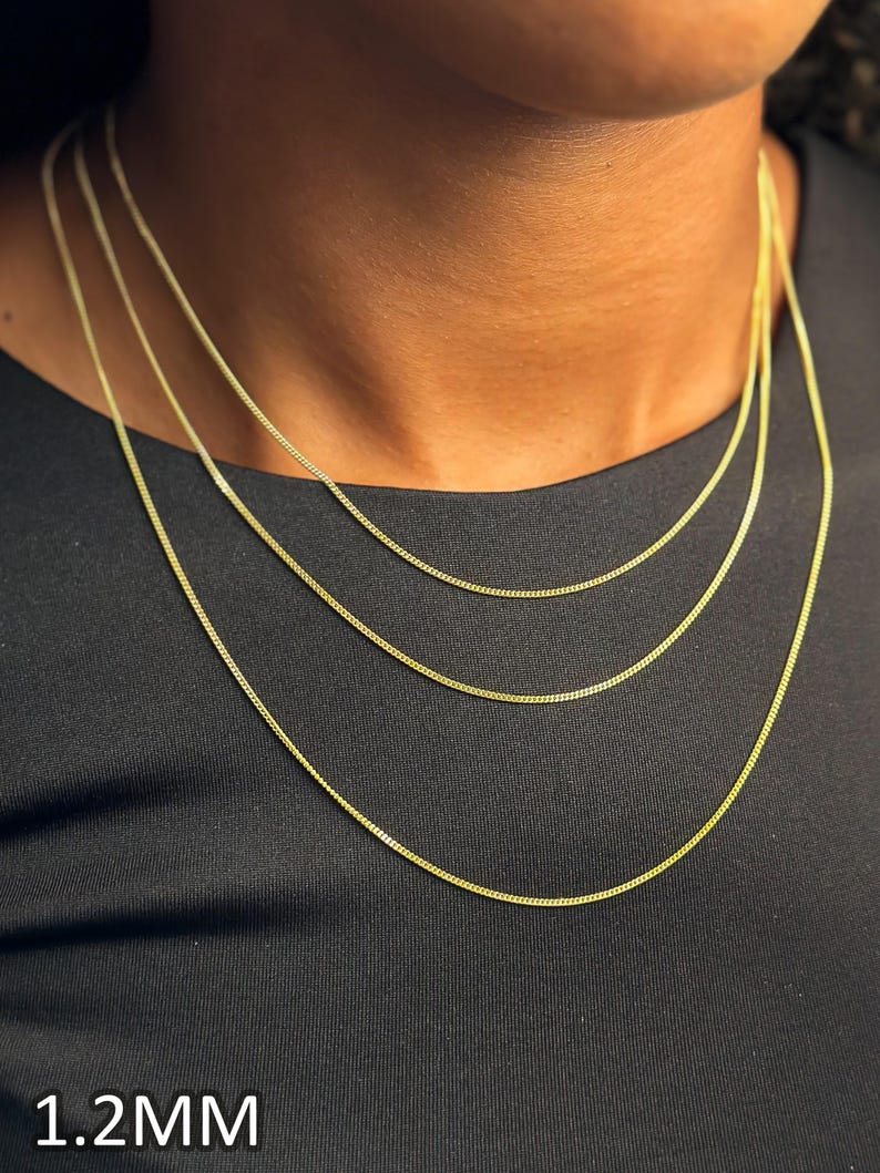 May include: Three layered gold-tone chains with a delicate curb link design. The chains are worn around the neck, featuring a consistent 1.2mm width. The metallic finish is bright and reflective, creating a classic layered look against a dark fabric.
