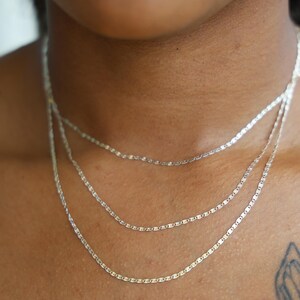 Puede incluir: Primer plano de tres delicados collares de plata superpuestos en un cuello. Cada collar presenta una fina cadena con peque&ntilde;os eslabones planos y rectangulares. Los collares son de diferentes longitudes, creando un efecto de capas.