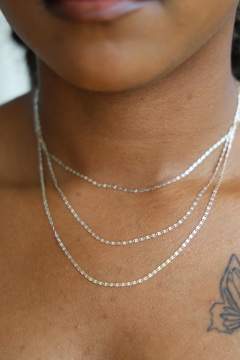 Puede incluir: Primer plano de tres delicados collares de plata superpuestos en un cuello. Cada collar presenta una fina cadena con peque&ntilde;os eslabones planos y rectangulares. Los collares son de diferentes longitudes, creando un efecto de capas.
