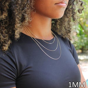 May include: A set of three thin gold-colored chain necklaces worn in a layered style. Each chain features a delicate link design and a polished finish. The text 1MM is clearly visible in the bottom right corner of the image, indicating the width.