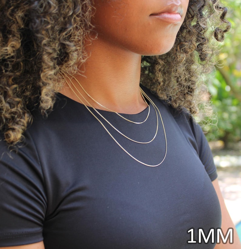 May include: A set of three thin gold-colored chain necklaces worn in a layered style. Each chain features a delicate link design and a polished finish. The text 1MM is clearly visible in the bottom right corner of the image, indicating the width.