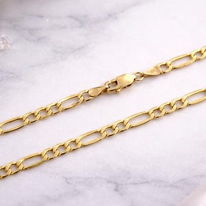 Op de afbeelding: Een goudkleurige Figaro-ketting. De ketting heeft een patroon van langwerpige ovale schakels afgewisseld met kortere, afgeronde schakels. De sluiting is ook goudkleurig. De ketting wordt gepresenteerd op een wit en grijs marmeren oppervlak.