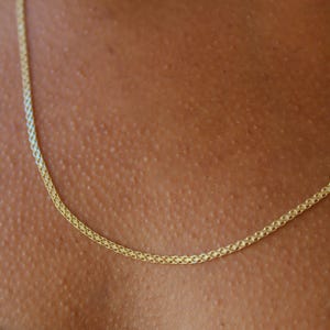 May include: A delicate gold chain necklace featuring a fine, woven wheat link design. The metallic yellow gold links are tightly interconnected, creating a smooth, continuous texture that rests against the skin in a simple, elegant circular drape.