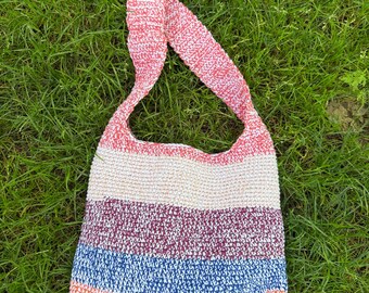 Könnte beinhalten: Handgefertigte Häkelschultertasche mit Streifenmuster. Die Tasche hat einen rosa-weißen Riemen, und der Körper der Tasche hat horizontale Streifen in Creme, Lila, Blau, Orange und Grün. Die Tasche liegt auf einer grünen Wiese.