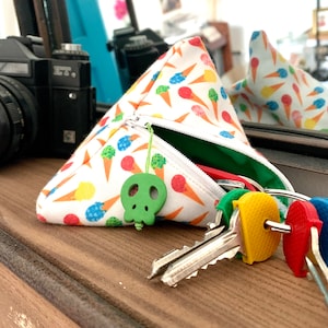 Puede incluir: Una bolsa triangular blanca con cremallera, estampada con coloridos conos de helado. Un dije verde con forma de calavera cuelga de la cremallera. Se ven llaves con cubiertas de colores.