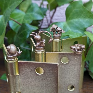 May include: A close-up of a decorative brass hinge with miniature bronze sculptures of a snail, mouse, mushrooms, and a frog. The hinge has a rectangular shape with circular cutouts. Green leaves are in the background.
