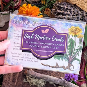 May include: A package of "Herb Medica Cards" featuring a dandelion illustration. The purple label reads "16 Herbal Property Cards, Double Sided 4" x 6". The background has handwritten text and a watercolor effect. The package is held by a hand.