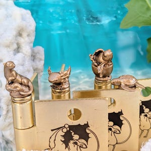 May include: A collection of brass-colored bottle stoppers featuring animal designs, including an otter, axolotl, turtle, and a creature emerging from an egg. The stoppers are displayed in front of gold-colored tags with black illustrations.