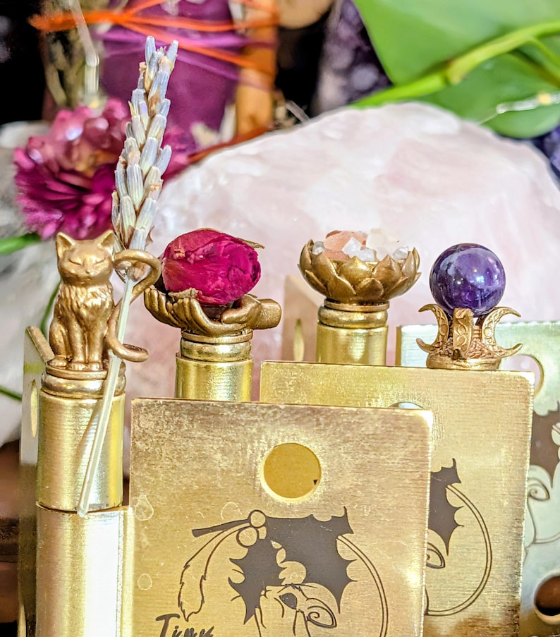 Four 3D-printed witch hinge heads in gold finish, each holding a magical element: lavender sprig, dried rosebud, crystal salts, and amethyst bead, set against a pink quartz crystal backdrop. Together the elements make a self-love spell