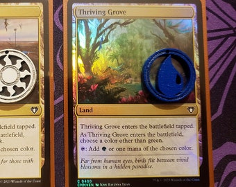 Może przedstawiać: Dwie karty Magic: The Gathering, Thriving Moor i Thriving Grove, z odpowiadającymi im symbolami many. Thriving Moor to karta terenu z białym symbolem słońca. Thriving Grove to karta terenu z niebieskim symbolem łzy.