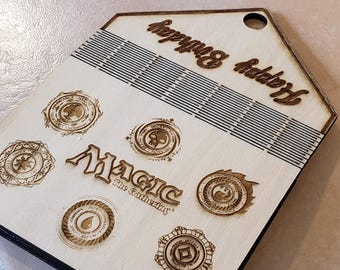 Może przedstawiać: Drewniane pudełko do przechowywania kart Magic: The Gathering z siatką do organizowania kart. Pudełko jest brązowe i zawiera logo Magic: The Gathering oraz kilka symboli.
