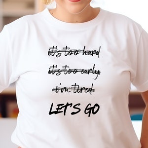K&ouml;nnte beinhalten: Wei&szlig;es T-Shirt mit schwarzem Text: "It's too hard, It's too early, I'm tired, LET'S GO." Der Text ist handschriftlich. Das T-Shirt hat kurze &Auml;rmel.