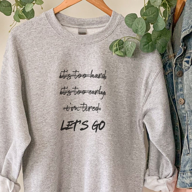 K&ouml;nnte beinhalten: Ein hellgraues Sweatshirt mit dem schwarzen Text "It's too hard, it's too early, I'm tired, LET'S GO". Das Sweatshirt h&auml;ngt an einem Holzb&uuml;gel. Auf der rechten Seite des Bildes ist eine Jeansjacke zu sehen.