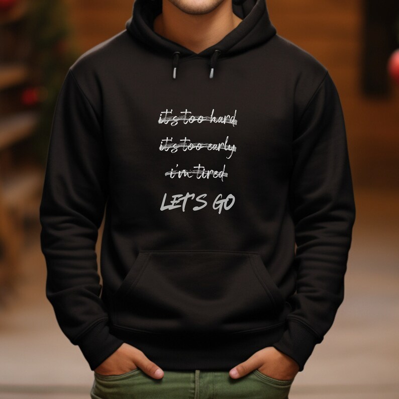 K&ouml;nnte beinhalten: Schwarzer Kapuzenpullover mit Fronttasche und Kordelzugkapuze. Der Text "it's too hard, it's too early, i'm tired, LET'S GO" ist in Wei&szlig; in einem handschriftlichen Stil gedruckt.