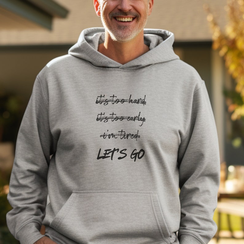 K&ouml;nnte beinhalten: Ein hellgrauer Kapuzenpullover mit Kordelzug und Fronttasche. Der Kapuzenpullover zeigt den Text "It's too hard, it's too early, I'm tired, LET'S GO" in schwarzer Schrift. Die Person, die den Kapuzenpullover tr&auml;gt, l&auml;chelt.
