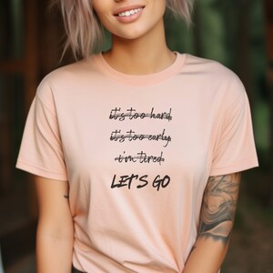 K&ouml;nnte beinhalten: Hellpfirsichfarbenes T-Shirt mit dem schwarzen Text "it's too hard, it's too early, i'm tired, LET'S GO". Das Shirt hat kurze &Auml;rmel und einen Rundhalsausschnitt.