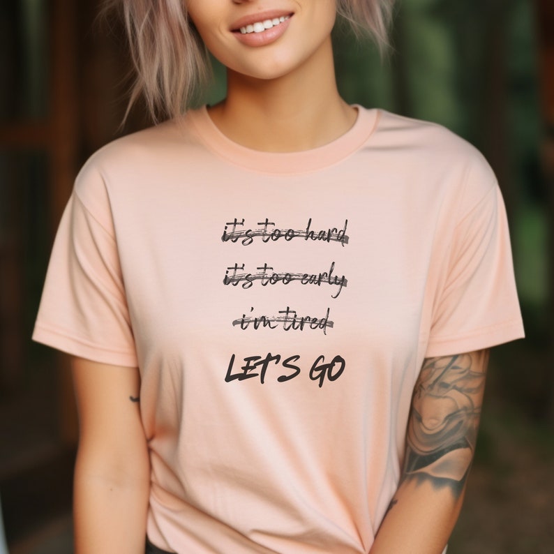 K&ouml;nnte beinhalten: Hellpfirsichfarbenes T-Shirt mit dem schwarzen Text "it's too hard, it's too early, i'm tired, LET'S GO". Das Shirt hat kurze &Auml;rmel und einen Rundhalsausschnitt.