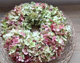 May include: A circular wreath constructed from dried hydrangea flowers featuring a color palette of soft pale green and muted pink petals, arranged in a dense, layered formation on a woven, circular natural fiber base.