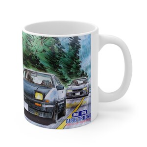 Op de afbeelding: Witte keramische mok met een aquarel illustratie van twee auto's die racen op een kronkelende weg. De auto's zijn lichtblauw en donkergrijs. De mok heeft een wit handvat en een blauwe en gele weg.