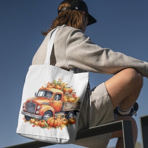 Pode incluir: Uma bolsa tote branca com um caminh&atilde;o vintage carregando ab&oacute;boras e folhas de outono. O caminh&atilde;o &eacute; laranja e vermelho com uma cabine branca. A bolsa est&aacute; sendo carregada por uma pessoa vestindo uma camiseta cinza e shorts.