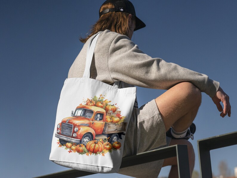 Pode incluir: Uma bolsa tote branca com um caminh&atilde;o vintage carregando ab&oacute;boras e folhas de outono. O caminh&atilde;o &eacute; laranja e vermelho com uma cabine branca. A bolsa est&aacute; sendo carregada por uma pessoa vestindo uma camiseta cinza e shorts.
