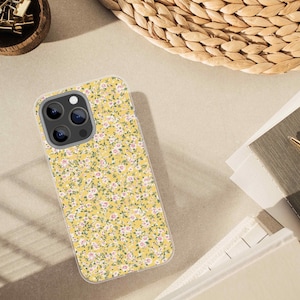 Mockup of a smartphone case printed with a yellow floral confetti pattern, placed on a beige desk beside a notepad, gold pen, and woven basket.