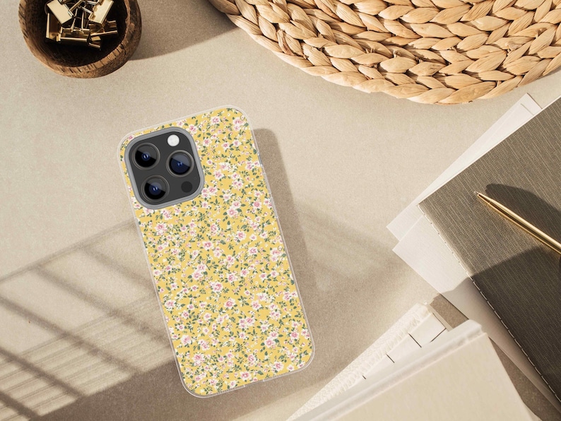 Mockup of a smartphone case printed with a yellow floral confetti pattern, placed on a beige desk beside a notepad, gold pen, and woven basket.