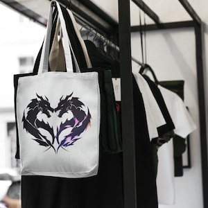 May include: White tote bag with black silhouette of two dragons facing each other in a heart shape. The bag has two handles and is hanging on a rack with other bags.