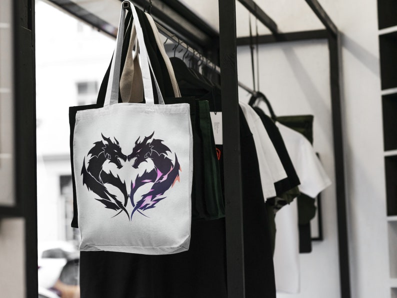 May include: White tote bag with black silhouette of two dragons facing each other in a heart shape. The bag has two handles and is hanging on a rack with other bags.