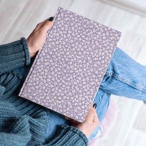 Close-up of a person holding a journal or sketchbook wrapped in a purple floral confetti pattern, seated cross-legged in a cozy indoor setting.