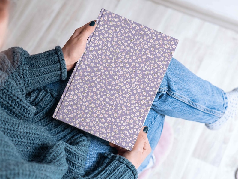 Close-up of a person holding a journal or sketchbook wrapped in a purple floral confetti pattern, seated cross-legged in a cozy indoor setting.