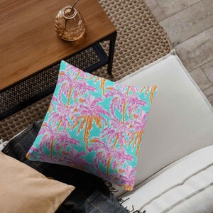 A throw pillow rests on a modern couch, covered in a seamless tropical pattern of pink and orange palm trees against a turquoise background, adding a vibrant summer touch to home decor.