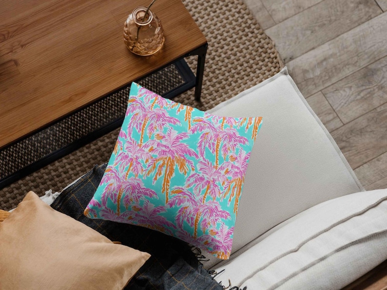 A throw pillow rests on a modern couch, covered in a seamless tropical pattern of pink and orange palm trees against a turquoise background, adding a vibrant summer touch to home decor.