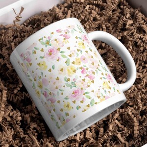 A white ceramic mug wrapped in a pastel floral pattern featuring pink and yellow roses with golden heart accents, displayed in a gift box with brown shredded paper, perfect for customized spring-themed gifts.
