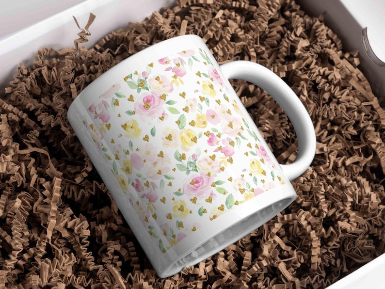 A white ceramic mug wrapped in a pastel floral pattern featuring pink and yellow roses with golden heart accents, displayed in a gift box with brown shredded paper, perfect for customized spring-themed gifts.