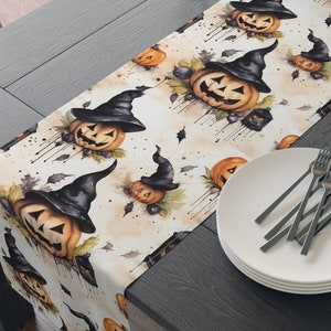 May include: A white table runner with a Halloween theme featuring a repeating pattern of orange pumpkins with black witch hats. The pumpkins are painted in a watercolour style with dripping paint accents. The runner is laid on a dark wood table with a plate and cutlery.