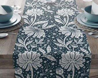 Puede incluir: Un camino de mesa con estampado floral blanco sobre fondo azul oscuro. El camino de mesa está colocado sobre una mesa de madera con cubiertos a cada lado.