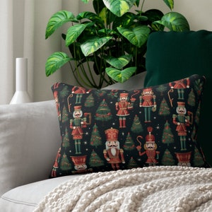 Op de afbeelding: Een zwart en groen kussen met een herhalend patroon van notenkrakerfiguren en kerstboompjes. Het kussen ligt op een witte bank met een groene deken.