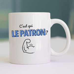 May include: White ceramic mug with a black outline of a bicep flexing with the text "C'est qui LE PATRON?" in blue letters.