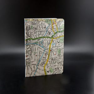 May include: A small, white notebook with a vintage map of Carshalton, England printed on the cover. The map features streets, parks, and landmarks in yellow, green, and blue.