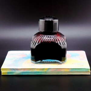 May include: A glass bottle of dark red ink with a black lid, sitting on top of a stack of colorful paper.