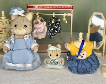 May include: A calico cat wearing a blue dress and a hat stands in front of a wooden clothing rack with hangers and a mirror. There are two other calico cats wearing dresses and a small yellow purse. The rack has a sign that says "Country Boutique".