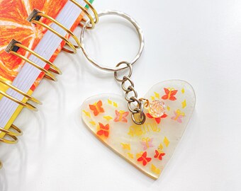 May include: A clear heart-shaped keychain with a silver chain and ring. The heart is decorated with small orange, yellow, and pink butterflies and the word "June" written in yellow.